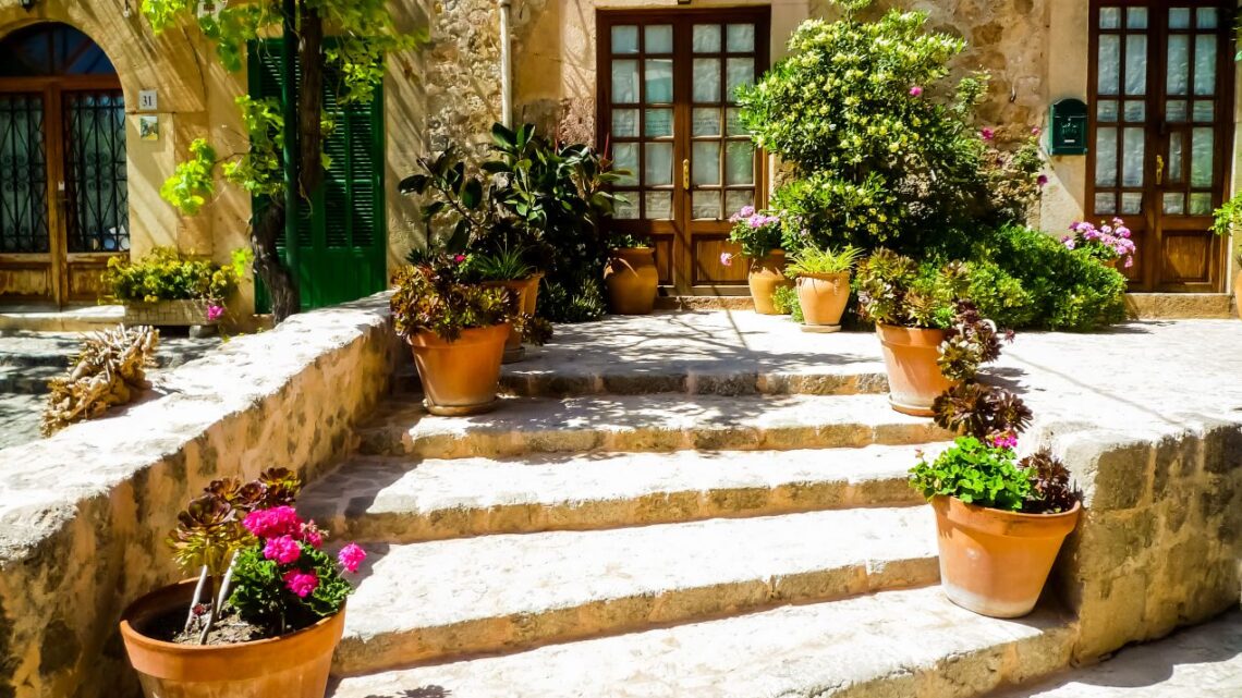 Mediterrane Steintreppe mit Terrakottatoepfen und Palettenkissen vor rustikalem Landhaus im Sueden