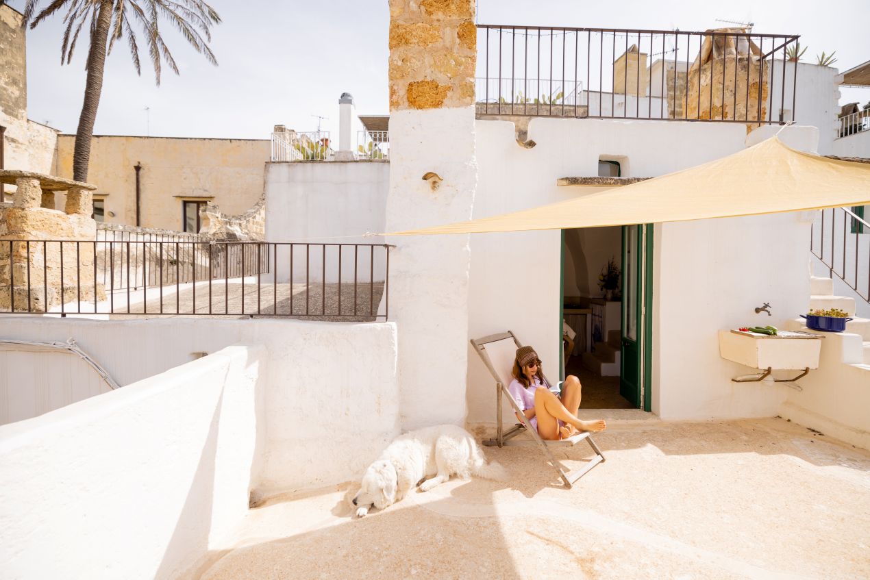 Frau sitzt auf Liegestuhl unter Sonnensegel vor mediterranem Haus mit Palettenkissen auf heller Dachterrasse